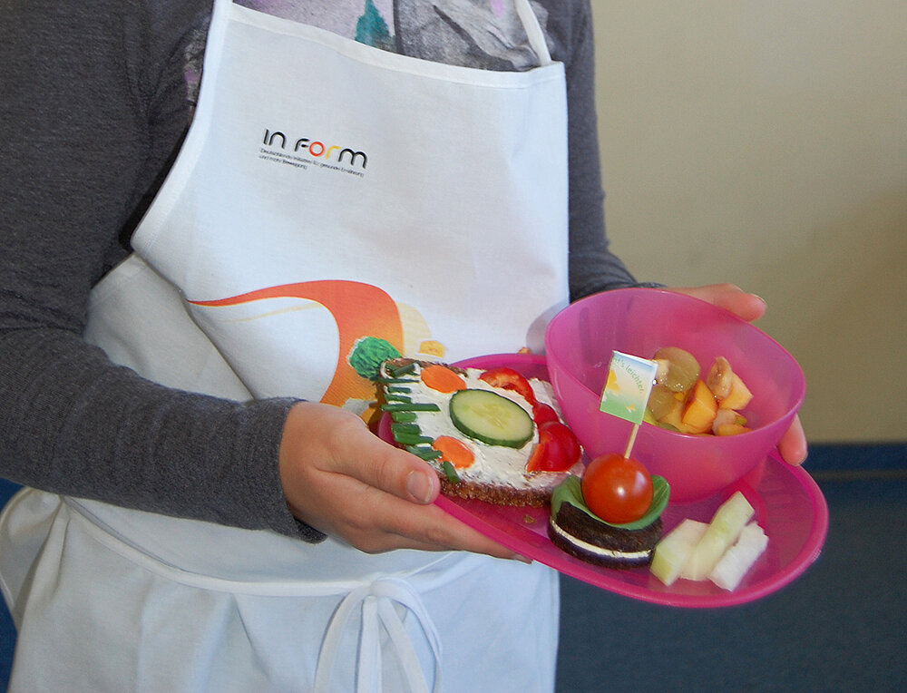 Kita- und Schulverpflegung Ein Mädchen hält einen Teller mit einem Brotgesicht und verschiedenem Obst und Gemüse in der Hand.