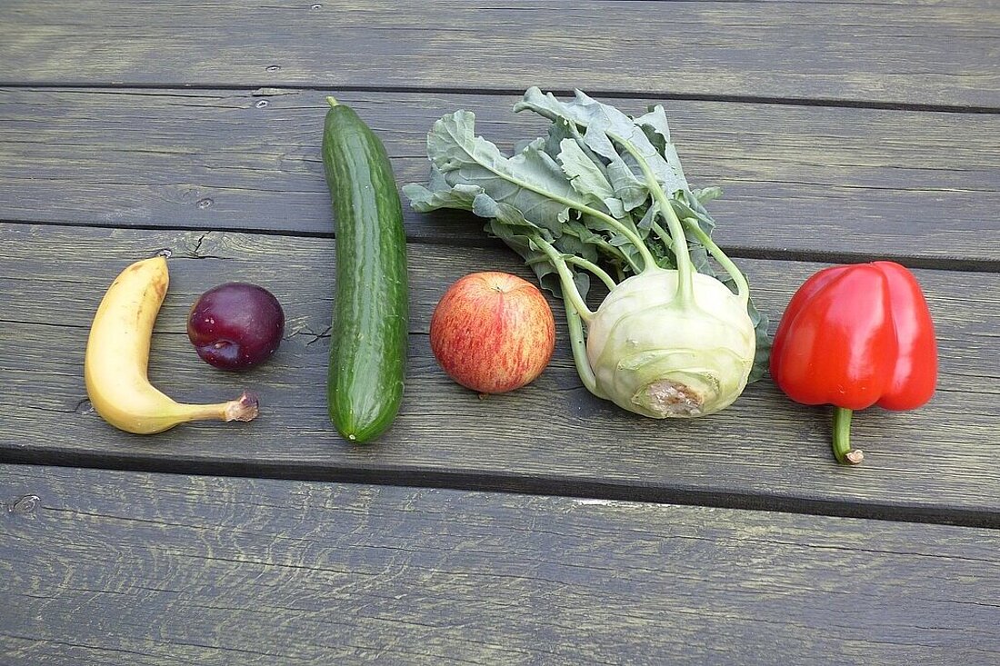 verschiedene Obst- und Gemüsesorten liegen in einer Reihe auf einen grünen Holztisch (1 Banane, 1 Pflaume, 1 Gurke, 1 Apfel, 1 Kohlrabi, 1 rote Paprika)