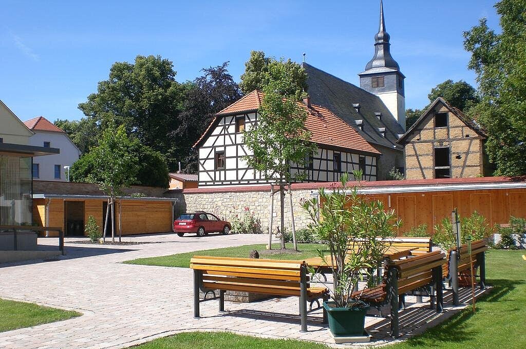 Gepflasteter Innenhof mit Bank, eingefasst von einer Wiese und Natursteinmauer. Im Hintergrund Fachwerkhäuser und eine Kirche.
